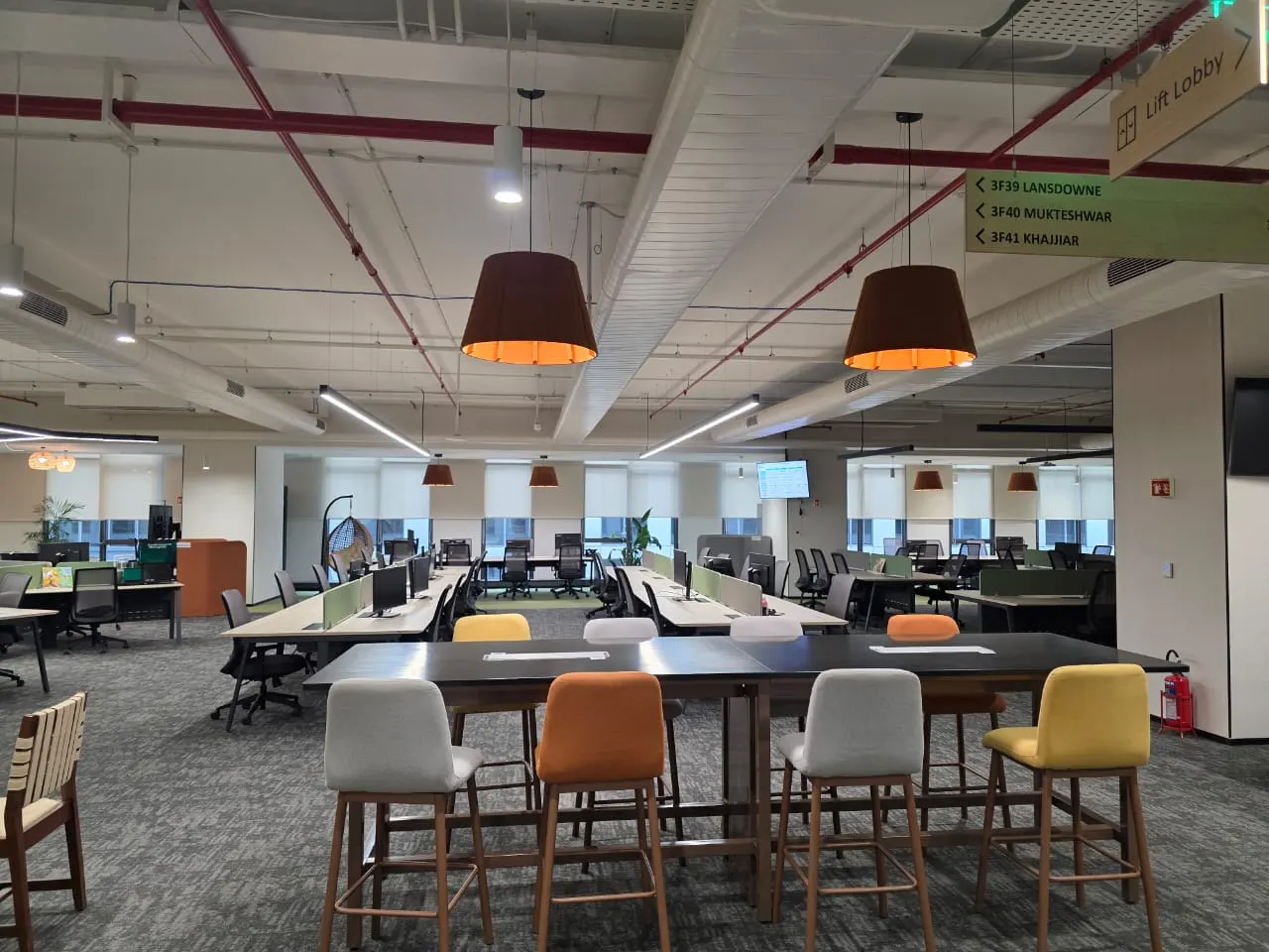 Open-plan office with large drum pendants over a collaboration table and linear lights beyond